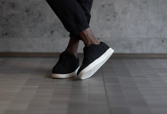 Person trägt schwarze Turnschuhe mit weißen Sohlen und steht mit überkreuzten Beinen auf gefliestem Boden.